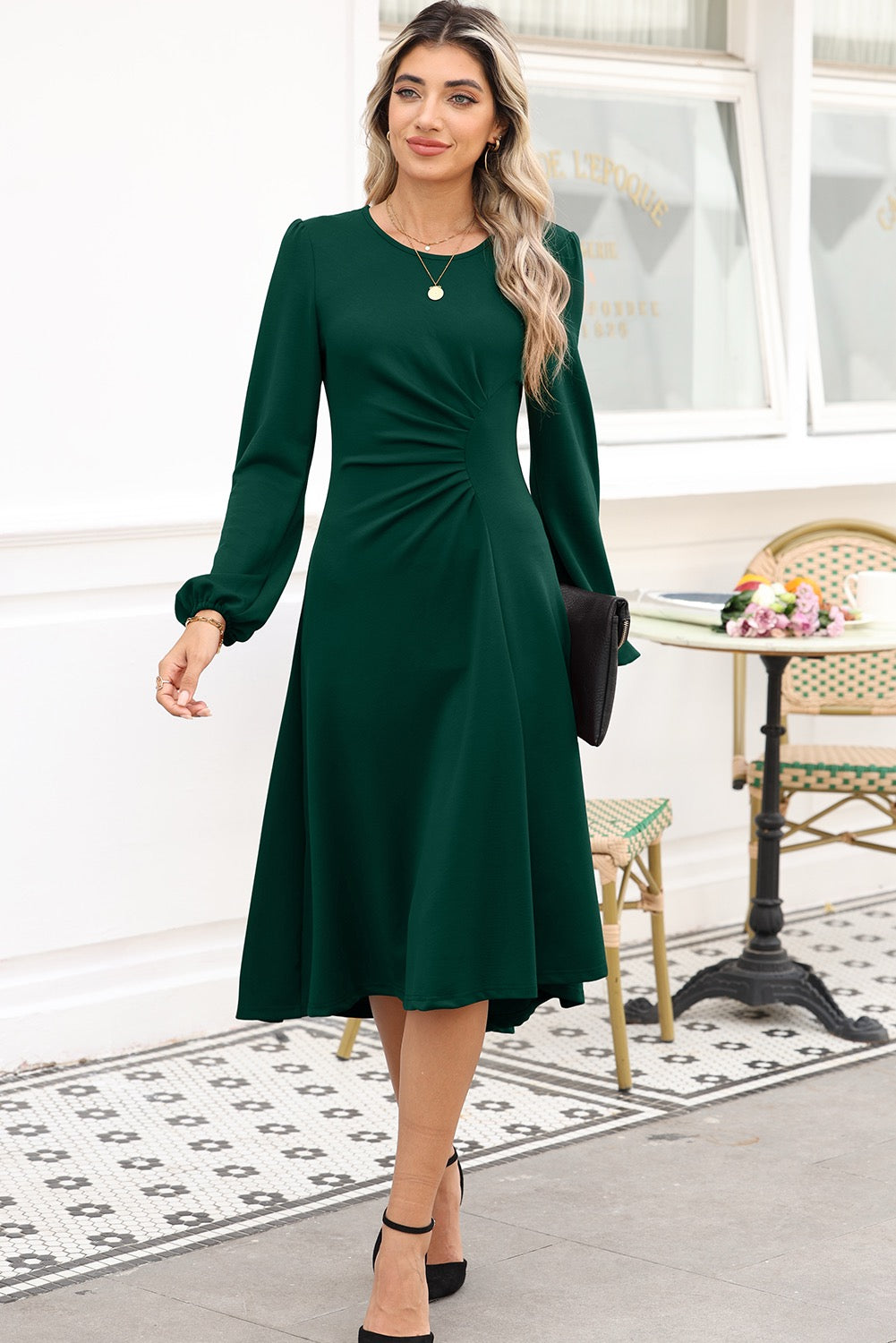 Woman wearing a green dress standing in an outdoor setting with a table and chairs in the background.